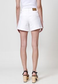 Pantalones cortos de mezclilla blanca con dobladillo deshilachado, cintura alta y bolsillos traseros. Una etiqueta de cuero marrón está cosida en la cinturilla. Sandalias de tacón negras puestas.