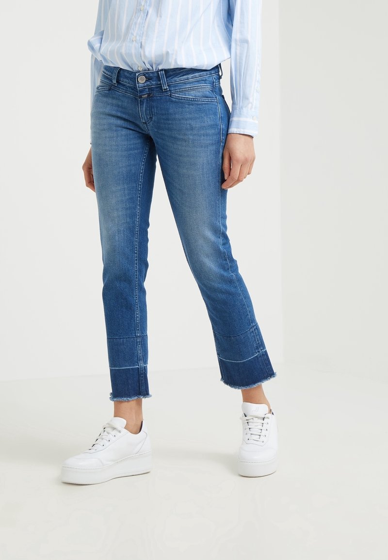 Jeans de mezclilla azul con un diseño recortado, dobladillo deshilachado y costuras en contraste. Llevados con zapatillas de plataforma blancas.