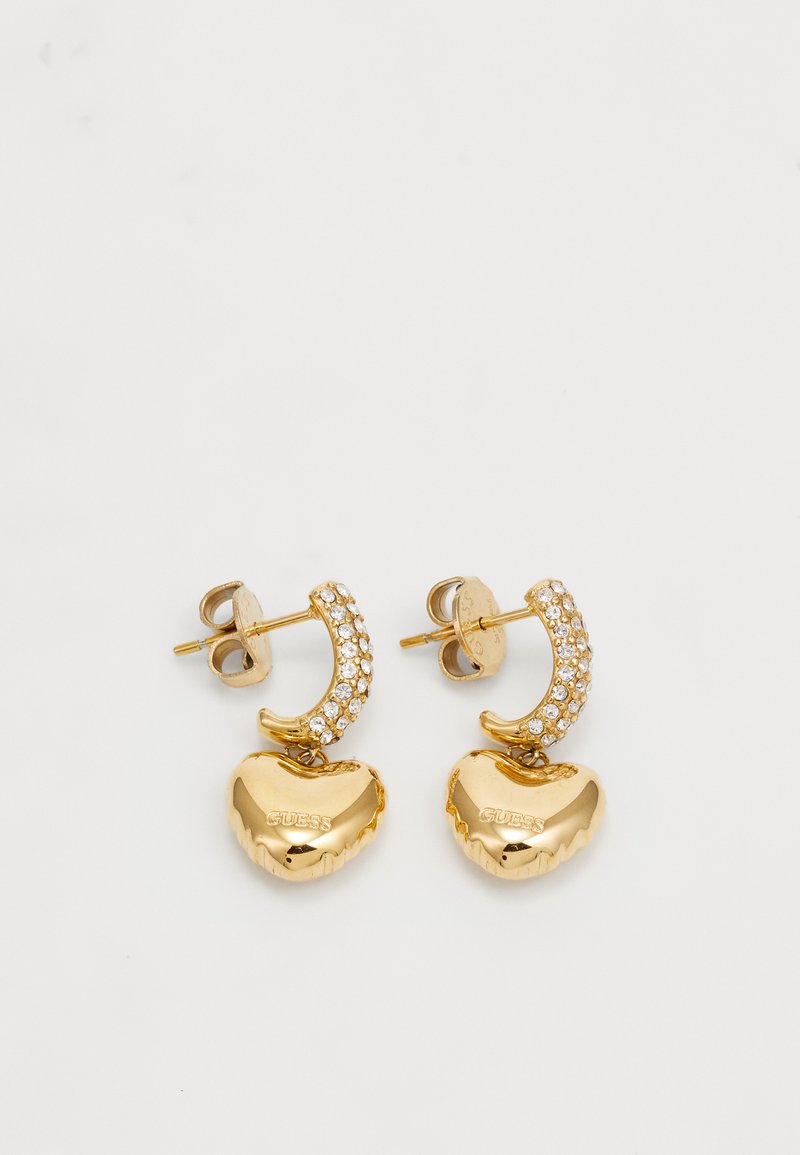 Boucles d'oreilles en ton doré avec des pendentifs en forme de cœur et des accents en rhinestone clair. Chaque boucle d'oreille présente un design de créole courbée. Marque visible.