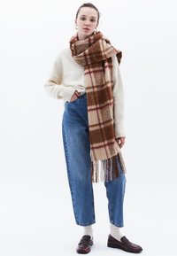 Karamell- och brunt rutigt scarf i mjukt tyg, med fransar i ändarna. Kombineras med en krämfärgad tröja och högmidjade blå jeans.