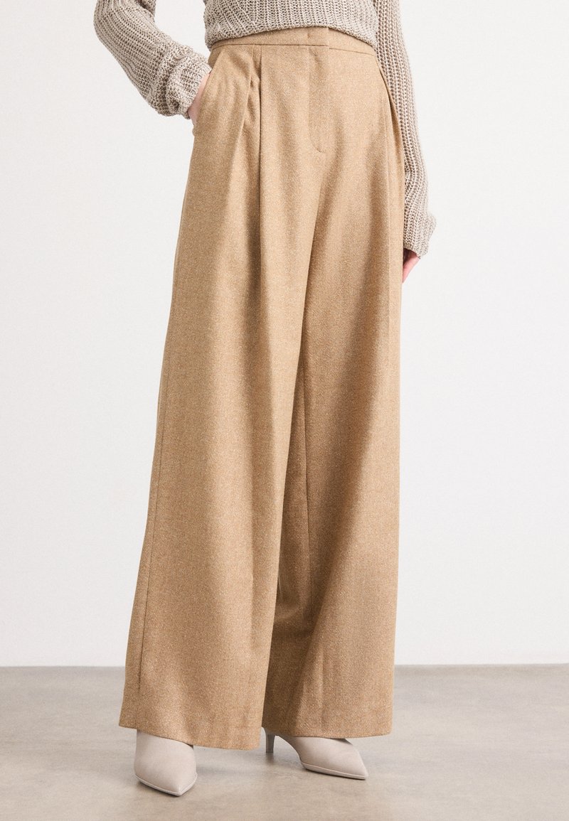 Beige széles szárú nadrág pliszőkkel, texturált anyagból, világosszürke hegyes orrú magassarkú cipővel párosítva. Minimalista design letisztult vonalakkal.