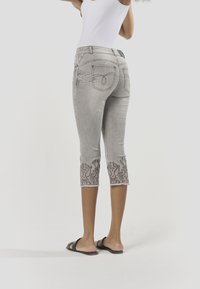 Szürke, rövidített farmernadrág részletesen hímzett szegéllyel. Testhezálló sziluettet, hátsó zsebeket és nyers szélű nadrágszárvégeket tartalmaz.