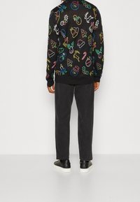 Person, der einen schwarzen Sweatshirt mit bunten Neon-Weihnachtssymbolen, dunklen Hosen und schwarzen Sneakers mit weißen Sohlen trägt, von hinten gezeigt.