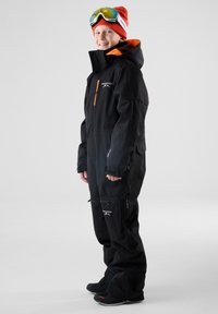 Zwart geïsoleerd skikostuum met oranje accenten, voorzien van een hoge kraag, ritszakken en versterkte manchetten. Model draagt skibril en muts.