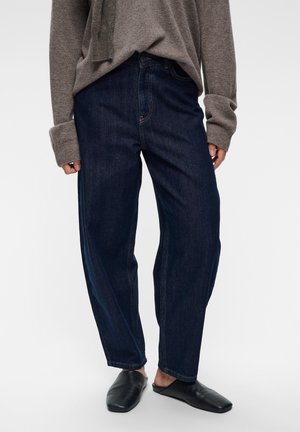Persoon draagt donkerblauwe loszittende jeans, grijze trui met lange mouwen en zwarte instapschoenen, staand op een lichte achtergrond.