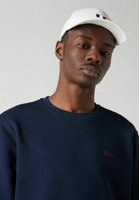 Donkerblauw sweatshirt met ribbeltextuur, voorzien van rode borduursel van "Parijs", gecombineerd met een witte pet met een blauw, wit en rode accent.