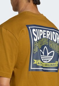 Camiseta de algodón marrón con una gran impresión gráfica azul que dice "SUPERIOR SPORTING GOODS" y presenta un logo de Adidas. Mangas cortas, cuello redondo.