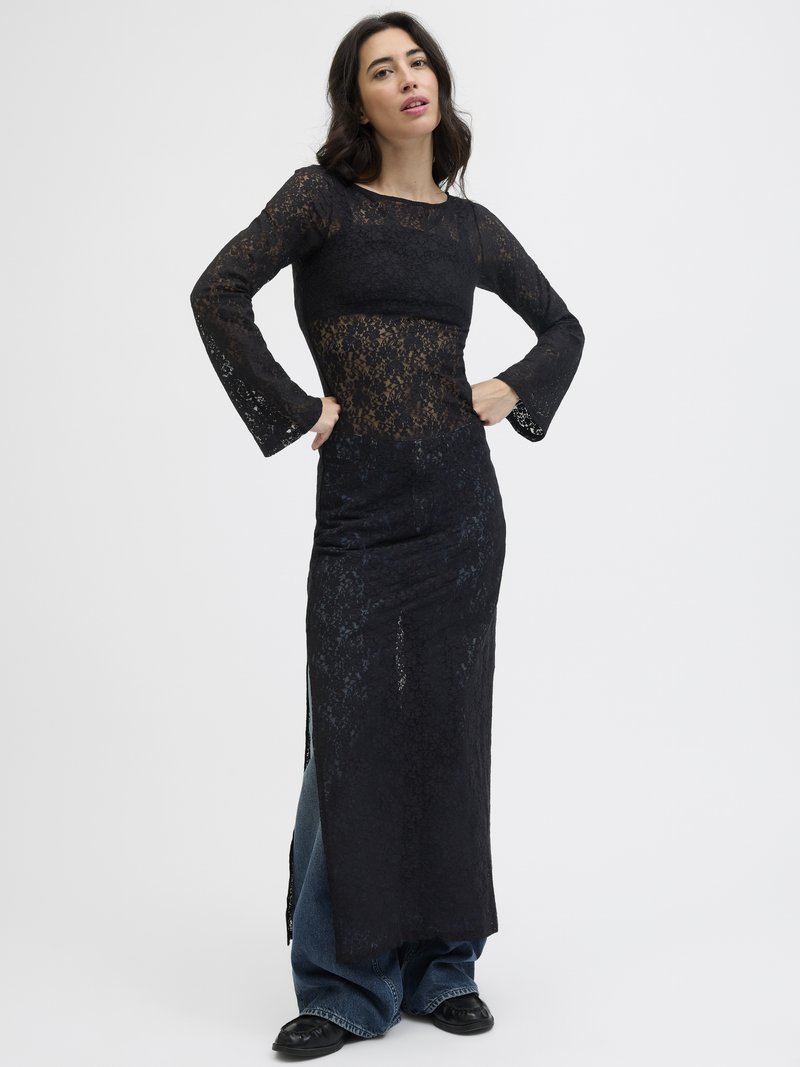 Vestido de renda preta com mangas longas e punhos amplos, apresentando uma sobreposição transparente e um design em camadas. Combinado com jeans de perna larga.