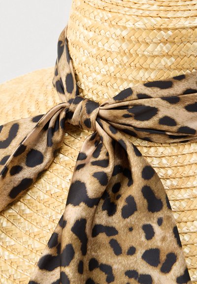 Φουλάρι με σχέδιο λεοπάρ, φτιαγμένο από απαλό ύφασμα, δεμένο στη βάση ενός πλεκτού ψάθινου καπέλου, με φυσικούς μπεζ τόνους και σκούρες κηλίδες.