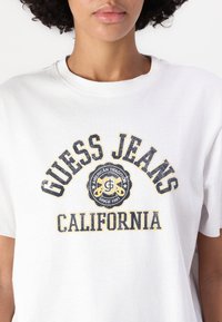 Bílý bavlněný tričko s grafickým potiskem s nápisy "GUESS JEANS" a "CALIFORNIA" v modré a žluté barvě, kulatým výstřihem a krátkými rukávy.