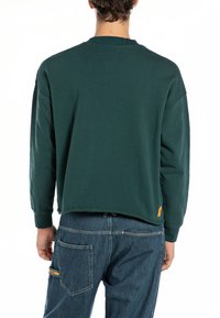 Felpa verde scuro a maniche lunghe con texture a coste, spalle cadenti e un piccolo etichetta arancione sull'orlo. La vista posteriore mostra una vestibilità rilassata.