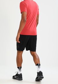 Mężczyzna ubrany w czerwoną koszulkę, czarne spodenki sportowe, białe skarpetki w wzory oraz czarne trampki Nike, stojący tyłem.