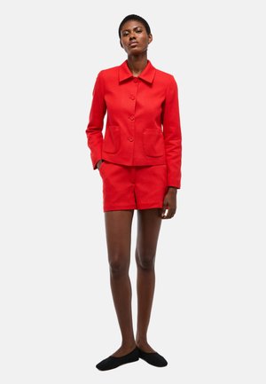 Femme debout portant une veste rouge boutonnée avec deux poches avant et un short rouge assorti, associé à des chaussures plates noires.