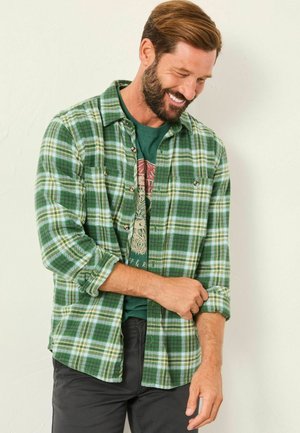 Grünes karierter Flanellhemd mit weißen und dunkelgrünen Akzenten, geknöpfte Vorderseite, hochgekrempelte Ärmel, getragen über einem dunkelgrünen Grafik-T-Shirt.