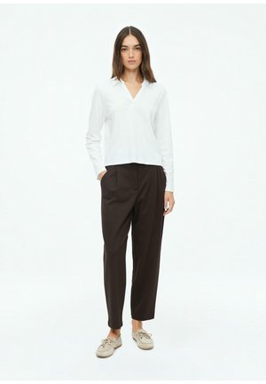 Frau steht, trägt ein weißes langärmeliges Hemd, schwarze hoch taillierte Hose und beige Schnürschuhe vor weißem Hintergrund.