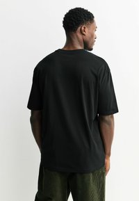 Tricou din bumbac negru cu o croială lejeră și mâneci scurte, având un design simplu, fără ornamente și un decolteu rotund standard.