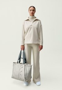 Moteris, vilkinti smėlio spalvos megztinį su aukštu kaklu ir atitinkančias kelnes, laiko didelę smėlio spalvos rankinę su tamsiai pilkais rankenėlėmis, stovinti prieš neutralų foną.