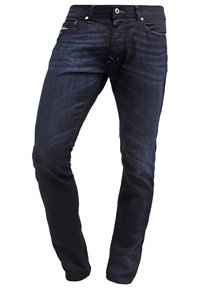 Jean en denim bleu foncé, coupe slim, fermeture à boutons sur le devant, poches avant et arrière, avec des détails d'usure subtils sur les cuisses.