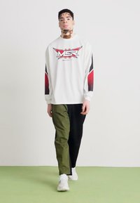Weißes Langarmhemd mit roten und schwarzen Akzenten, versehen mit einem Logo. Olivgrüne und schwarze Hose mit entspannter Passform. Weiße Sneaker.
