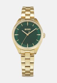 Montre-bracelet dorée avec un cadran vert, une lunette cannelée et des index horaires. Avec un bracelet en maillons et des aiguilles minimalistes ; le nom de la marque est affiché à 12 heures.