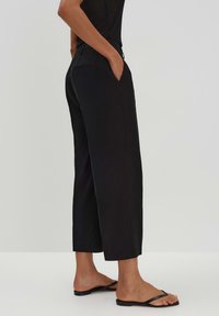 Black, high-waisted trousers with a straight cut, front pockets, and a slightly textured fabric. Worn with black flip-flops.
