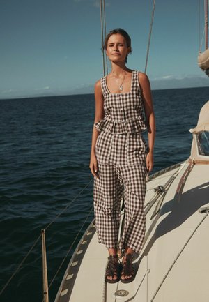 Young woman in brown checkered sleeveless top and pants standing on a sailboat deck with ocean in the background under clear sky.