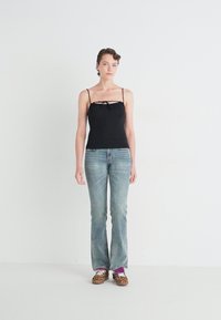 Schwarzes Spaghetti-Top mit einem Knoten-Detail vorne, kombiniert mit hellblauen Schlagjeans und Sneakers im Tierdruck. Weißer Hintergrund.