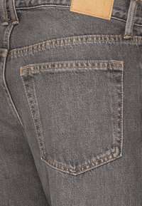 Primer plano de un bolsillo trasero de unos jeans de mezclilla gris con costura naranja y una etiqueta de cuero marrón en la cinturilla.