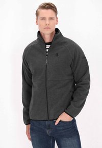 Giacca grigia con zip realizzata in materiale morbido, con colletto alto, presenta un logo sul petto e tasche laterali. Abbinata a jeans blu.