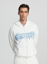 Felpa bianca con zip e logo blu consumato sul petto, caratterizzata da un tessuto morbido, cappuccio e tasche frontali. Texture liscia, vestibilità casual.