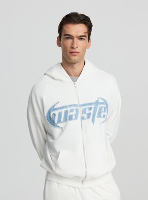 Felpa bianca con zip e logo blu consumato sul petto, caratterizzata da un tessuto morbido, cappuccio e tasche frontali. Texture liscia, vestibilità casual.