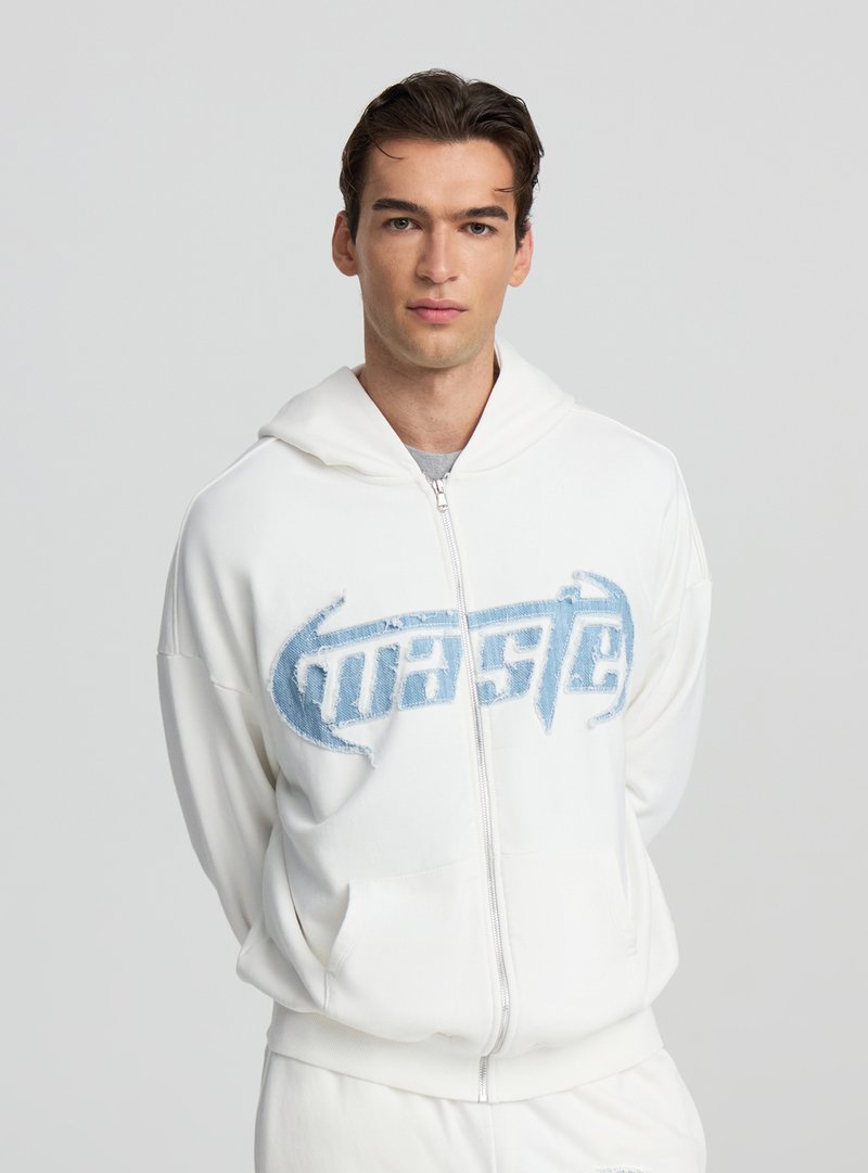 Felpa bianca con zip e logo blu consumato sul petto, caratterizzata da un tessuto morbido, cappuccio e tasche frontali. Texture liscia, vestibilità casual.