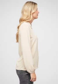 Blusa beige a maniche lunghe con polsini arricciati, scollatura tonda e tessuto morbido e dalla texture piacevole. Abbinata a jeans grigi strappati.