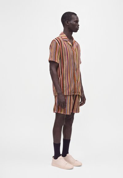 Homme debout de profil portant une chemise à manches courtes rayée multicolore et un short assorti avec des chaussettes noires et des baskets rose clair.