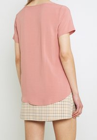 Miękki różowy top z krótkim rękawem i zaokrąglonym dołem, zestawiony z jasnobrązowo-kremową spódnicą w kratę. Gładka faktura i luźny krój.