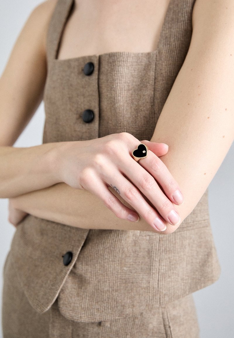 Anel em forma de coração com superfície em esmalte preto e borda em tom dourado, usado numa mão contra um fundo de tecido castanho texturizado.