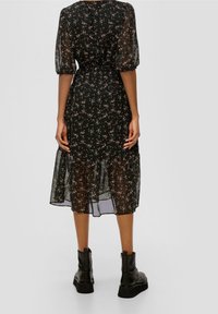 Kvinna i en svart blommig midikjol med genomskinliga puffärmar och nederkant, matchad med svarta platåankelstövlar, stående med avslappnade händer.