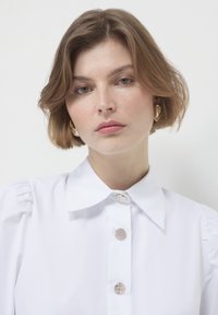 Young woman with short light brown hair wears gold hoop earrings and white blouse with decorative silver buttons, neutral background.