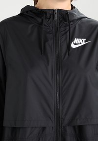 Musta Nike-tuulitakki hupulla, vetoketju koko edessä ja valkoinen Nike-logo vasemmassa rinnassa. Sileä, kevyt kangas.