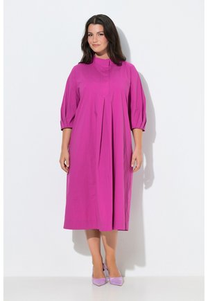 Frau trägt ein leuchtend pinkfarbenes Midikleid mit dreiviertel Ärmeln und Falten, steht vor einem weißen Hintergrund, trägt dazu passende pinke Absatzschuhe.