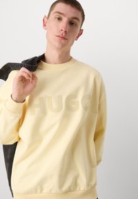 Sweatshirt de algodão amarelo com um grande logótipo "HUGO" em relevo na frente. O design tem um decote redondo e punhos canelados.
