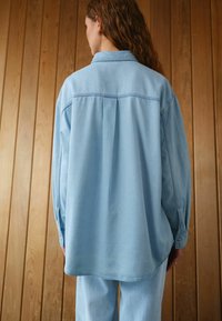 Lichtblauwe denim shirt met een ontspannen pasvorm, voorzien van een kraag, lange mouwen en een rugplooidetail. Gladde textuur.
