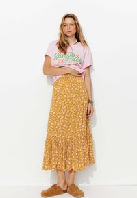 T-shirt graphique rose avec texte vert, associé à une jupe maxi florale jaune avec un ourlet volé, et des chaussures brunes à enfiler.