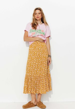 T-shirt graphique rose avec texte vert, associé à une jupe maxi florale jaune avec un ourlet volé, et des chaussures brunes à enfiler.
