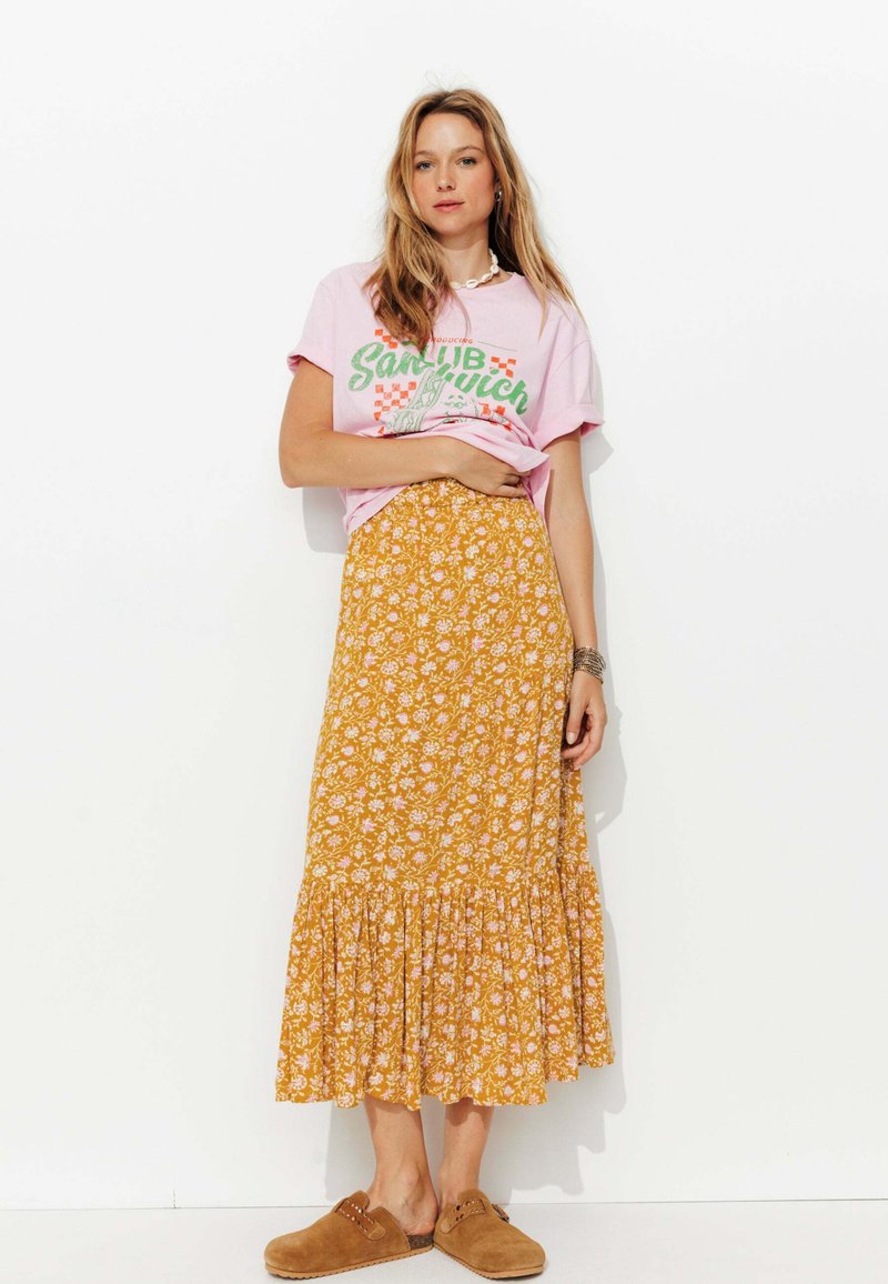 T-shirt graphique rose avec texte vert, associé à une jupe maxi florale jaune avec un ourlet volé, et des chaussures brunes à enfiler.