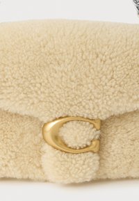 Krēmkrāsas, pūkaina tekstūra soma ar zelta metāla aizdari un noapaļotu aizvēršanas detaļu. Uzsvars uz materiāla mīkstumu un siltumu.