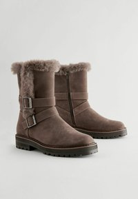Des bottines en daim marron présentent un revers en fausse fourrure, deux sangles à boucle, une fermeture éclair sur le côté et une semelle robuste pour une meilleure traction.