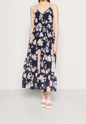 Femme portant une robe maxi sans manches bleu marine à fleurs avec un col en V et des sandales plateformes roses sur fond blanc uni.