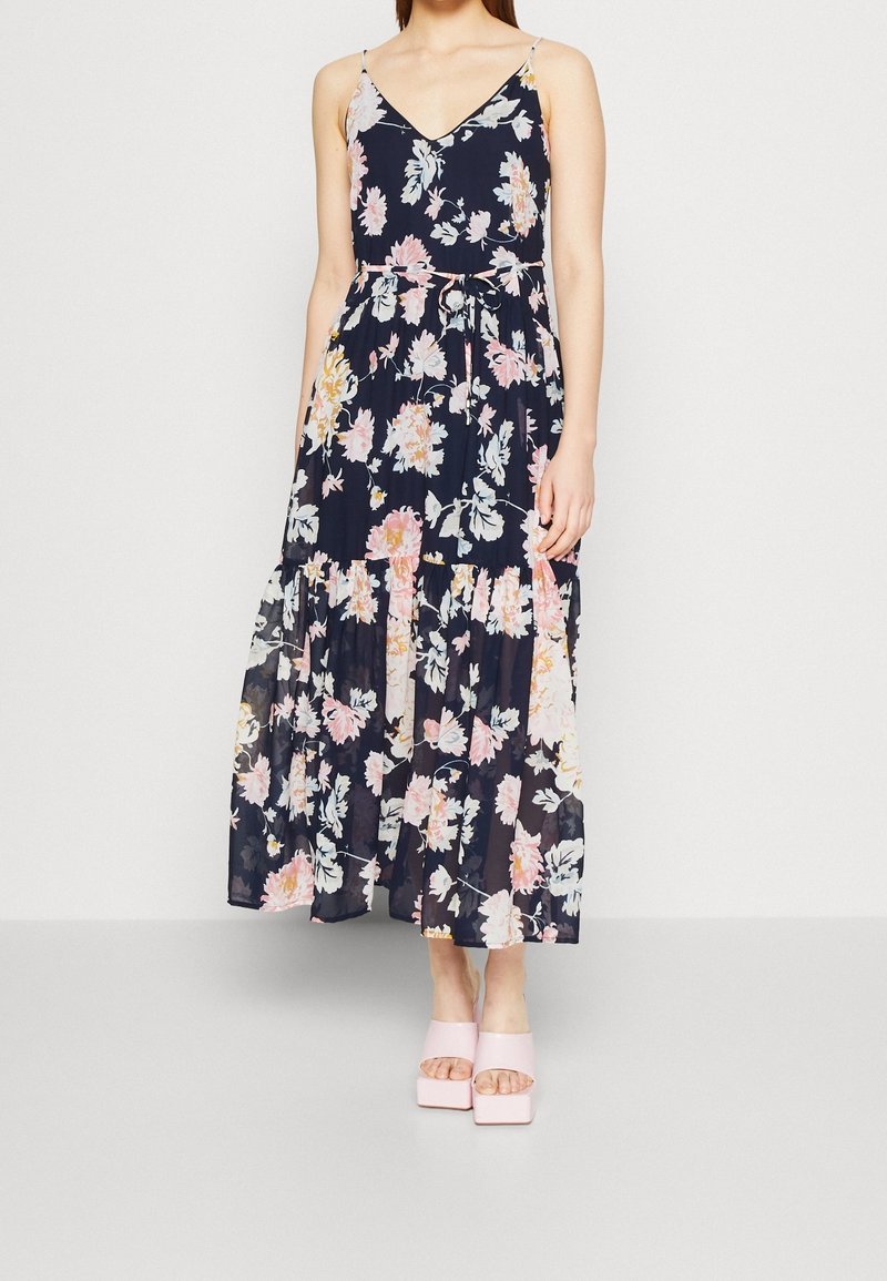 Femme portant une robe maxi sans manches bleu marine à fleurs avec un col en V et des sandales plateformes roses sur fond blanc uni.