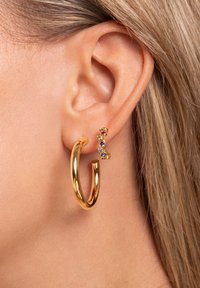 Orecchino a cerchio d'oro con finitura lucida accanto a un piccolo chiodo d'oro con pietre preziose colorate. L'orecchio è parzialmente visibile.